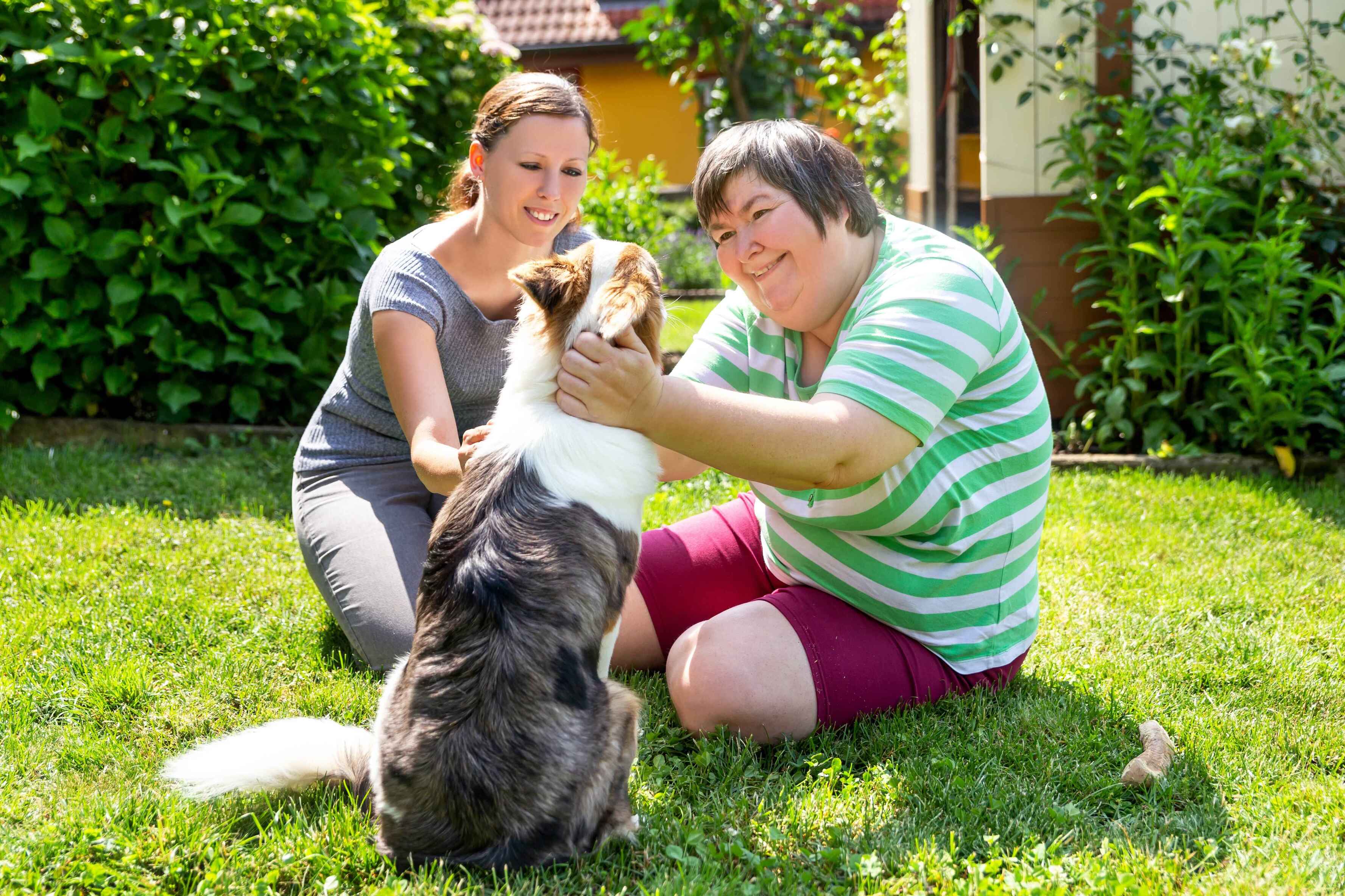 Two women, one with a disability, play with a dog on a lawn