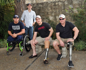 Photo: Three seated men pose for the camera. Two of them sit on a bench and have prosthetic legs. One sits in a wheelchair and has double leg amputations. A fourth man stands behind the group.