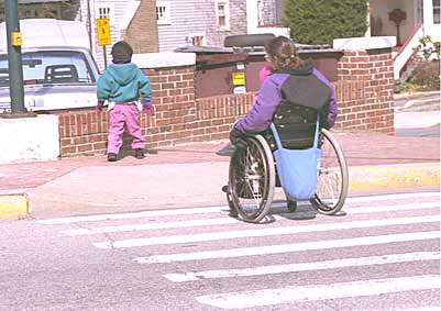A photograph of a woman using a wheelchair crossing the street at a crosswalk behind a young child.