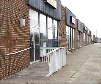A photograph of a one-story brick building with several store fronts, one of which has an accessible ramp leading to the entrance of a restaurant. The wooden ramp extends into the wide sidewalk area.
