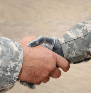 A close-up of a handshake between two service members in camouflage uniforms. One person's hand is a prosthesis.